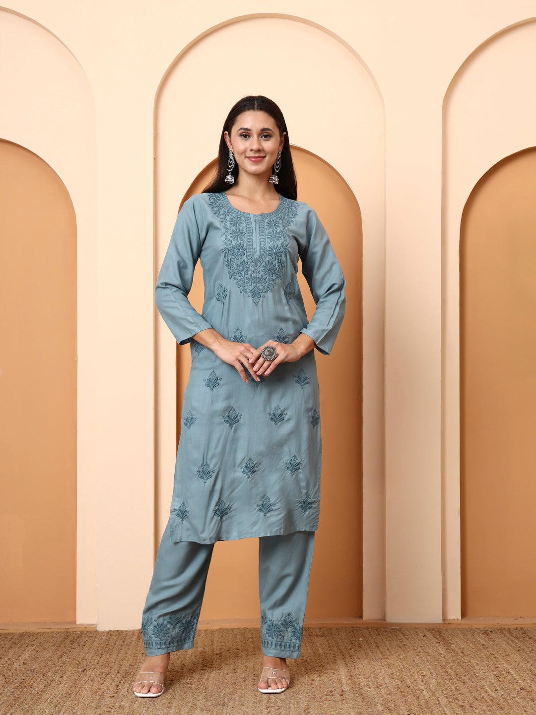 Woman wearing a light green traditional outfit with floral patterns against a beige wall.